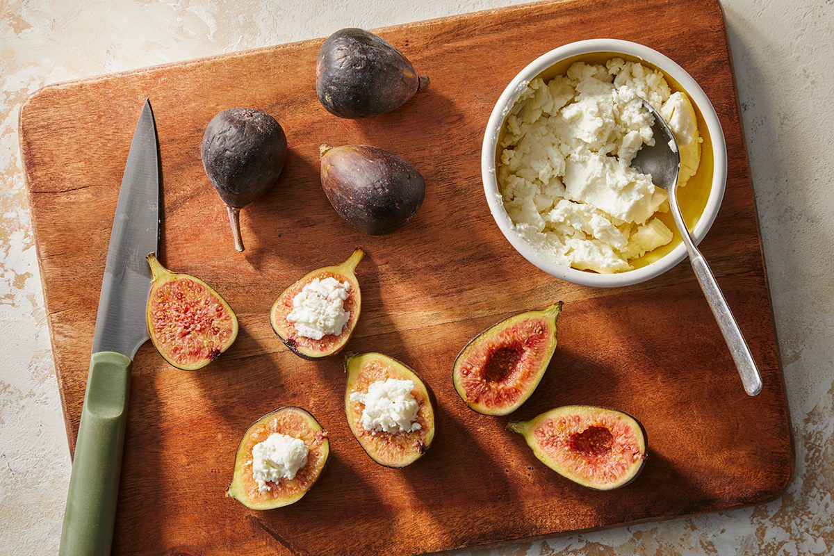 A wooden cutting board with fresh figs, some whole and some halved, a bowl of soft cheese with a spoon, and a knife. The halved figs are topped with cheese.