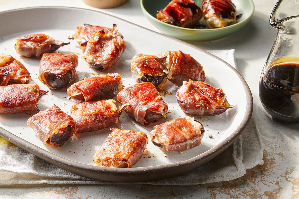 A white platter filled with bacon-wrapped dates, freshly baked and slightly crispy, sits on a table next to a small jug of dark sauce and a green bowl with additional servings in the background.