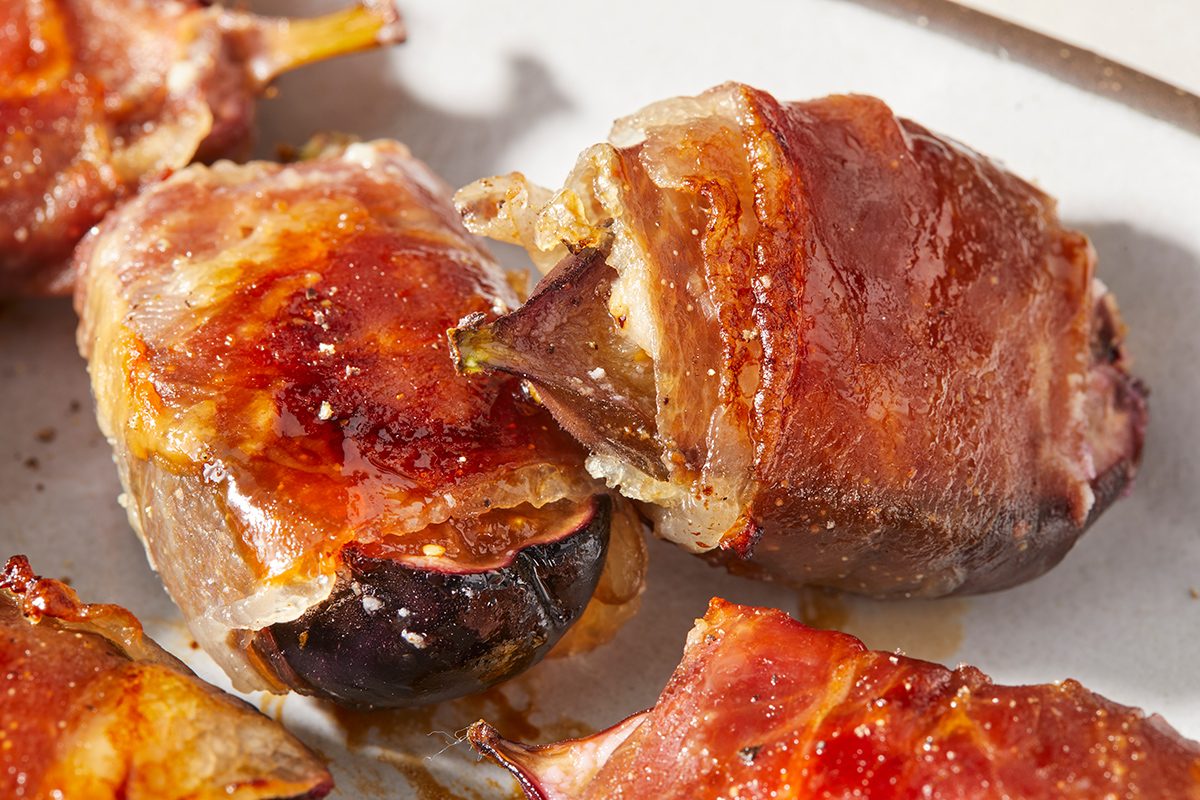Close-up of roasted figs wrapped in crispy prosciutto, served on a white plate. The figs have caramelized edges and the prosciutto appears golden and slightly glossy.