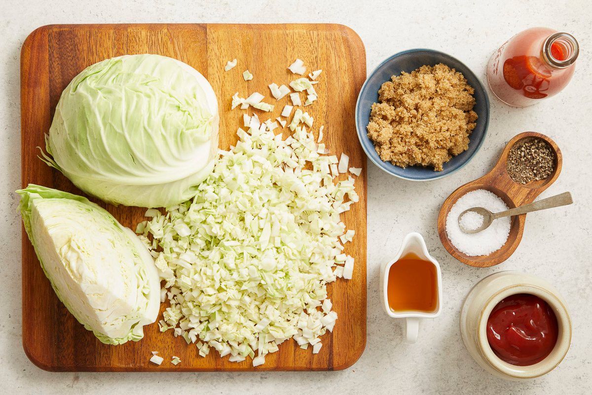 Overhead horizontal image of cabbage, brown sugar, ketchup, red sauce, cider vinegar, salt and pepper on a light gray countertop