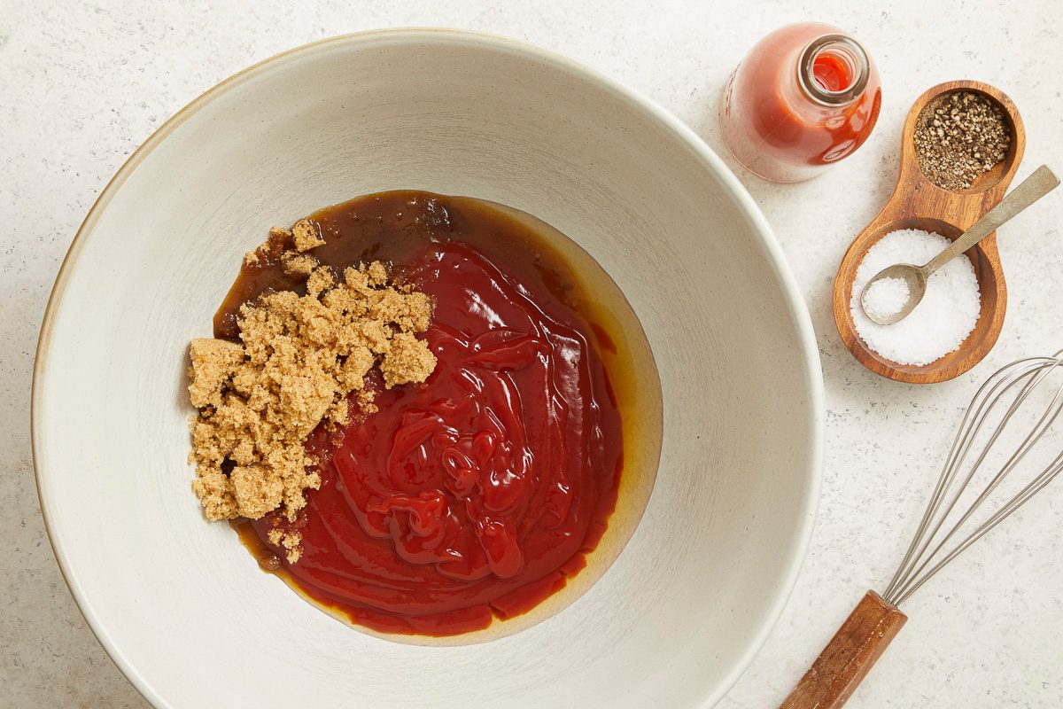 Overhead horizontal image of brown sugar, ketchup, and a dark brown sauce base being combined in a large white mixing bowl.