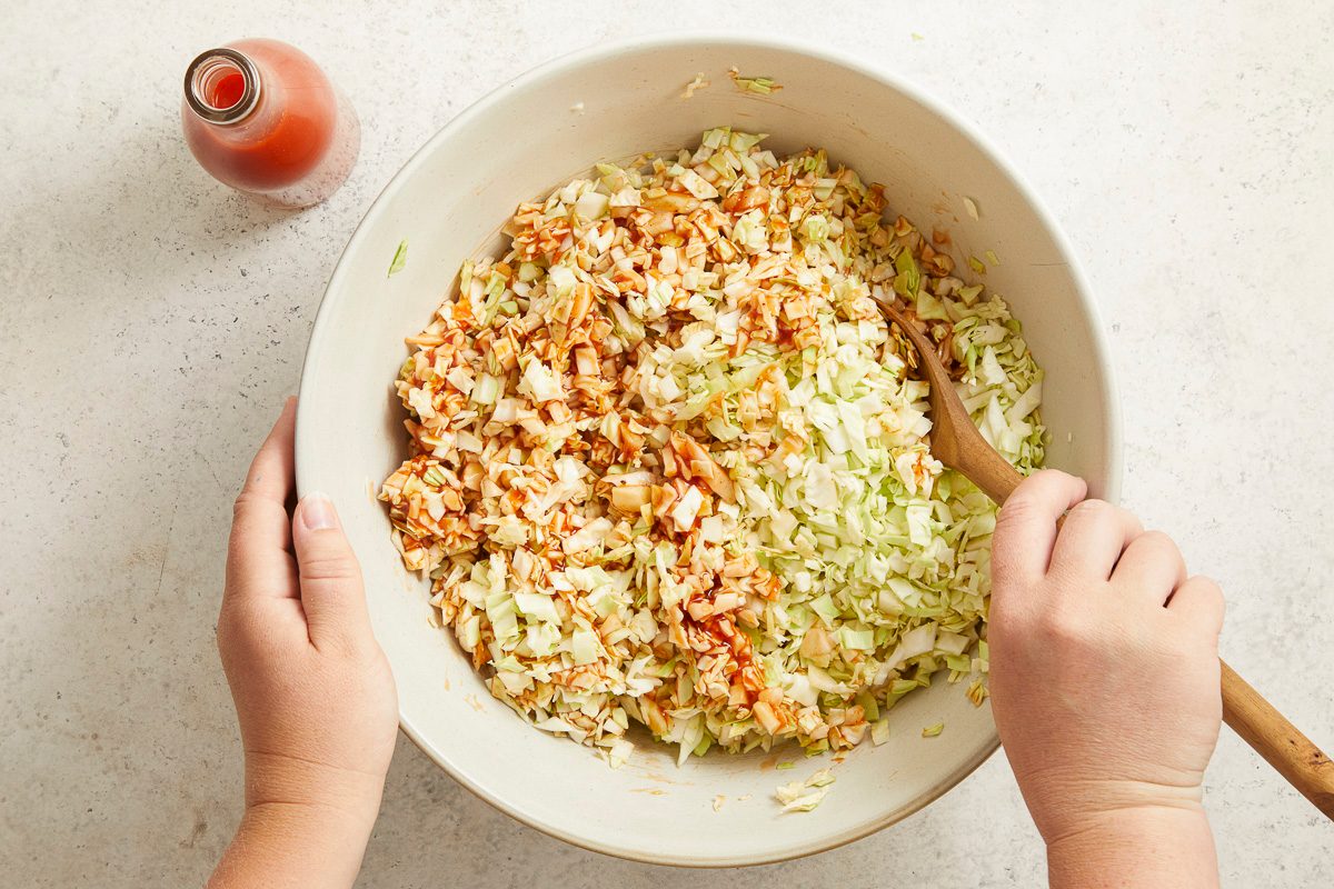 Overhead horizontal image of hands mixing shredded cabbage and a reddish-brown sauce in a large white bowl with a wooden spoon