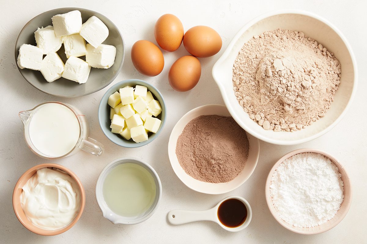 Baking ingredients on a white surface, including eggs, flour, cocoa powder, powdered sugar, cream cheese, butter, sour cream, buttermilk, and vanilla extract in separate bowls.