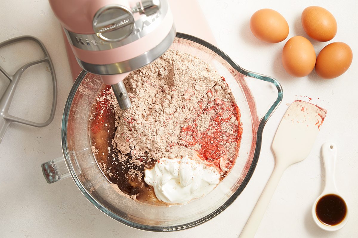 A stand mixer with a glass bowl is mixing cake ingredients, including dry mix, eggs, sour cream, and vanilla. Nearby are four whole eggs, a spatula, a metal beater, and a small dish of vanilla extract on a white surface.
