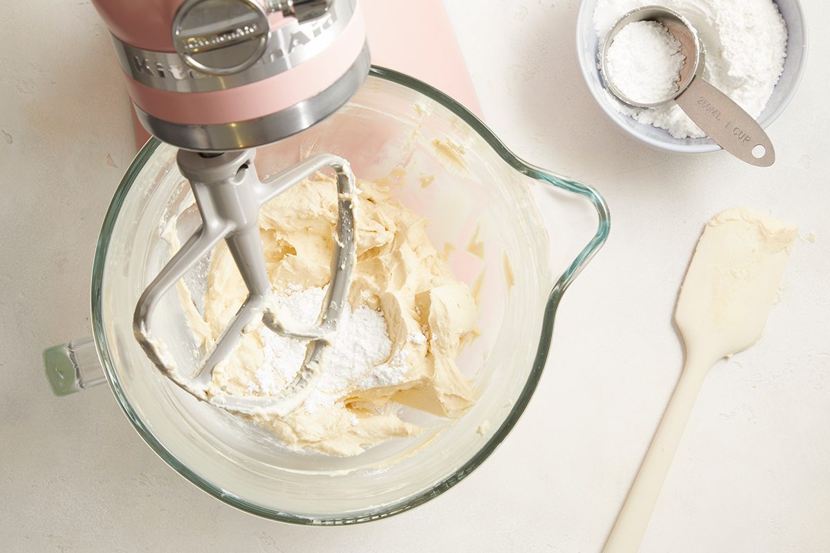 A stand mixer with a paddle attachment mixes a creamy batter in a glass bowl. Nearby are a white spatula and a small bowl of powdered sugar with a metal measuring cup.