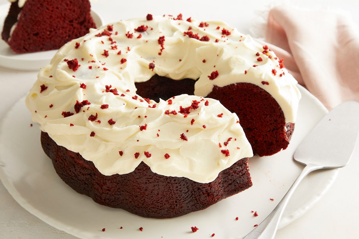 A red velvet bundt cake with cream cheese frosting and red velvet crumbs on top sits on a white plate. A slice has been cut and placed in the background, next to a cake server and a light pink napkin.
