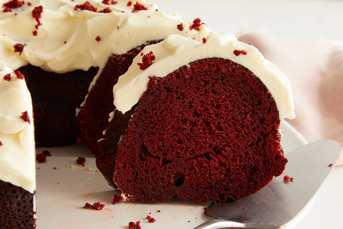 A close-up of a sliced red velvet bundt cake topped with creamy white frosting and red cake crumbs, with a cake server underneath a slice on a white plate.