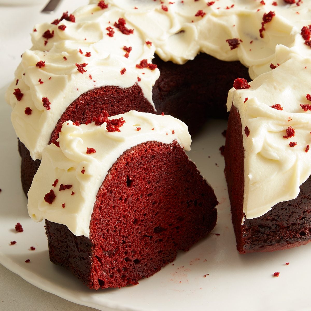 A red velvet bundt cake with a thick layer of white cream cheese frosting and red cake crumbs sprinkled on top, with one slice cut and slightly pulled out on a white plate.