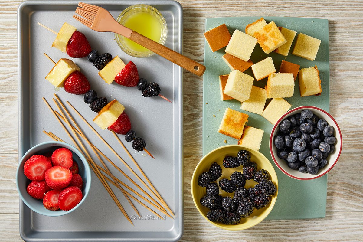 Overhead horizontal image of ingredients and partially assembled fruit and cake skewers. Cubes of cake, whole and sliced strawberries, blueberries, and blackberries are arranged on a baking sheet and a cutting board, with some threaded onto bamboo skewers alongside a small bowl of glaze and a pastry brush.