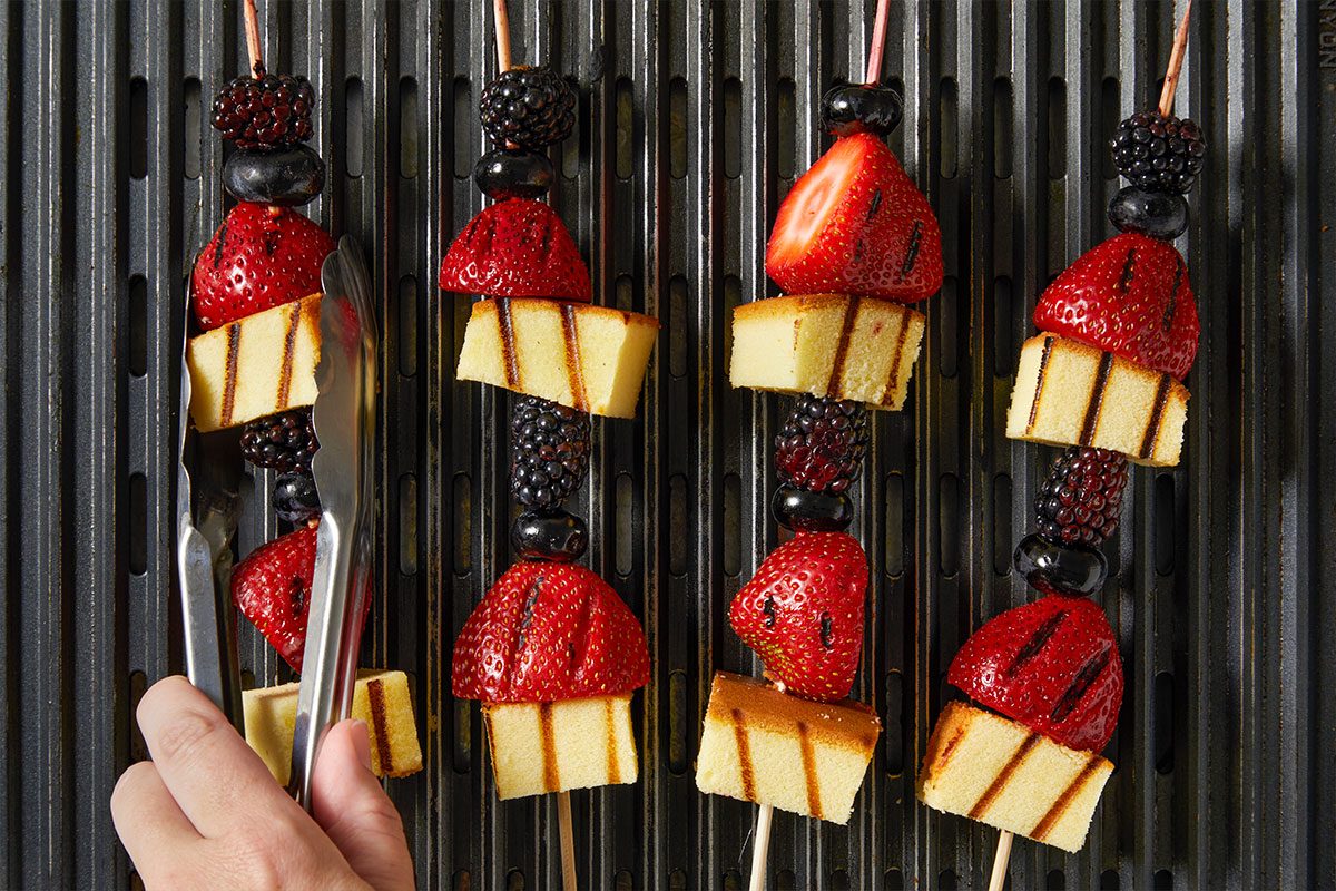 Overhead horizontal image of a hand using tongs to rotate a skewer on a grill pan. The skewer features pieces of pound cake, grilled strawberries, blueberries, and blackberries. Several other similar skewers are visible on the grill, all showing distinct grill marks.