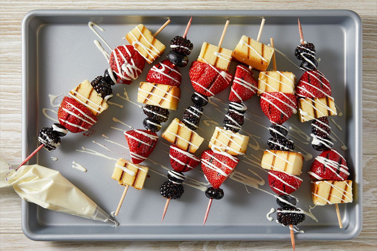 Overhead horizontal image of several dessert skewers arranged on a baking sheet. The skewers are loaded with grilled pound cake pieces, halved strawberries, blueberries, and blackberries, all generously drizzled with white icing. A piping bag filled with white icing is visible in the bottom left corner.