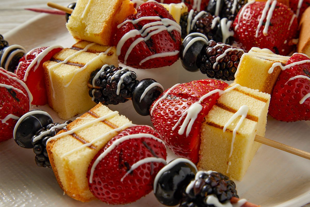 Eye Level horizontal image of multiple dessert skewers arranged on a white platter. Each skewer features grilled pound cake cubes, grilled strawberries, blackberries, and blueberries, all generously drizzled with white icing.