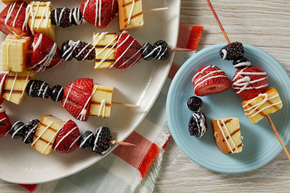 Overhead horizontal image of dessert skewers featuring cubes of grilled pound cake, fresh strawberries, blueberries, and blackberries, all drizzled with white icing. Several skewers are arranged on a white serving platter, with one individual skewer and additional berries and cake pieces on a small blue plate next to a checkered napkin.