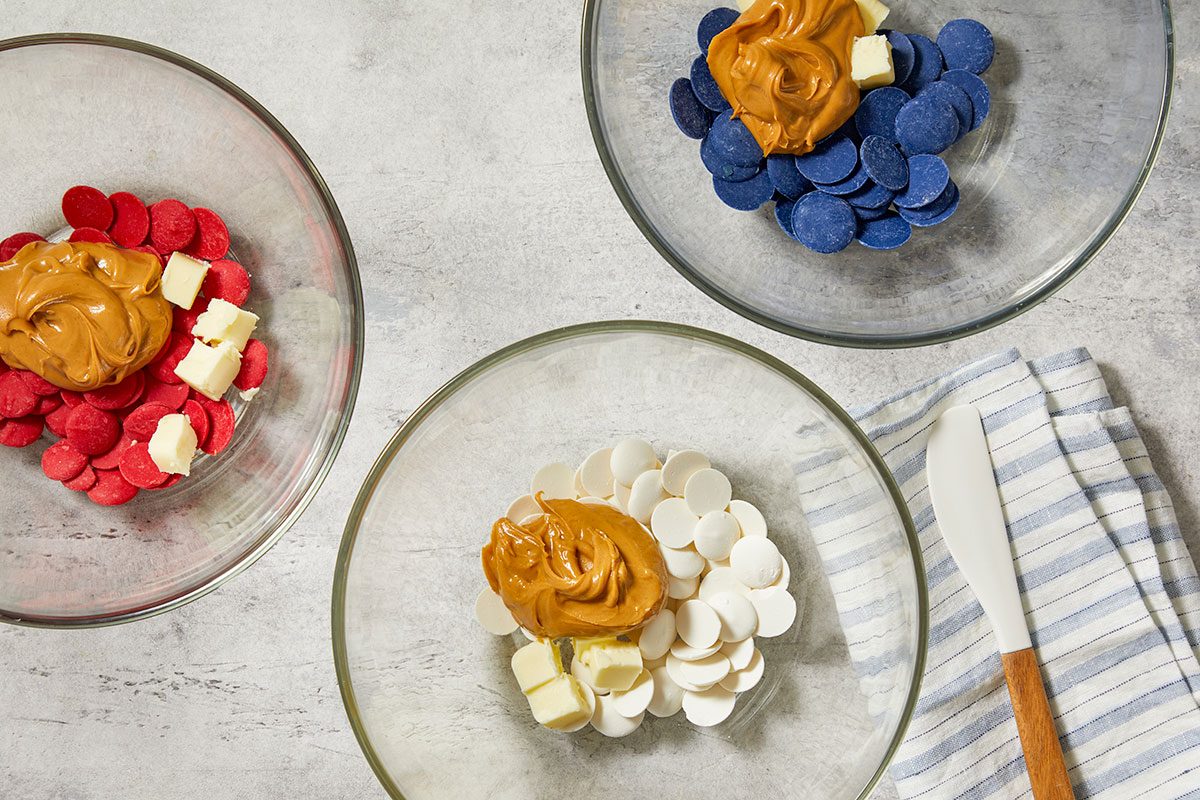 Overhead horizontal image of three clear glass bowls on a light grey background, each containing different colored melting wafers, a dollop of peanut butter, and cubes of butter. The bowls hold red, white, and blue melting wafers respectively, alongside peanut butter and butter, with a white spatula resting on a striped towel to the right.
