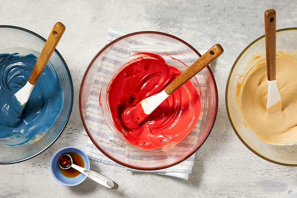 Overhead horizontal image of three glass bowls containing different colored mixtures. One bowl holds a vibrant red mixture with a white spatula, another contains a blue mixture with a spatula, and a third has a tan-colored mixture. A small blue bowl with a 1/2 teaspoon measuring spoon filled with a dark liquid is also visible next to a striped kitchen towel.