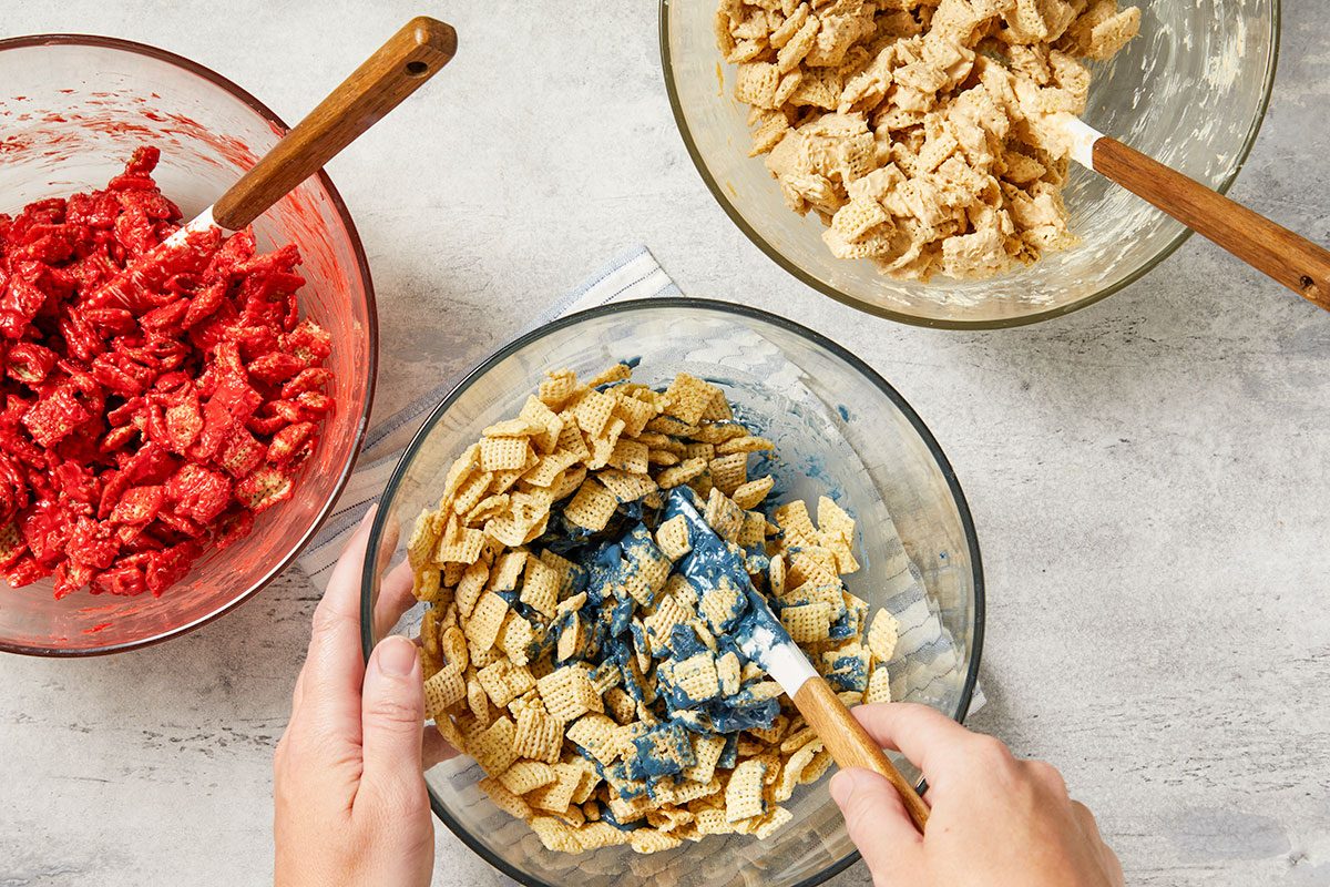 Overhead horizontal image of hands mixing cereal squares with a blue coating in a glass bowl, using a white spatula with a wooden handle. Two other glass bowls are visible: one filled with cereal squares coated in red, and another with cereal squares coated in a light, possibly white or tan, substance.