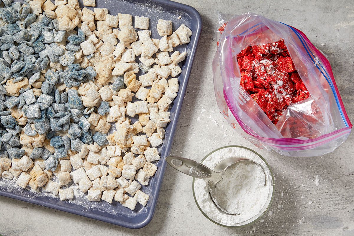 Overhead horizontal image of a baking sheet filled with light beige and blue powdered sugar-coated cereal pieces. Next to the sheet, a plastic bag holds red powdered sugar-coated cereal, alongside a bowl of powdered sugar with a measuring scoop.