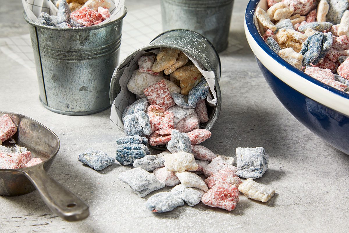 Eye Level horizontal image of red, white, and blue snack mix, heavily coated in powdered sugar, spilling from a tipped galvanized bucket onto a gray surface. Additional snack mix is visible in an upright bucket, a blue bowl, and a metal scoop.