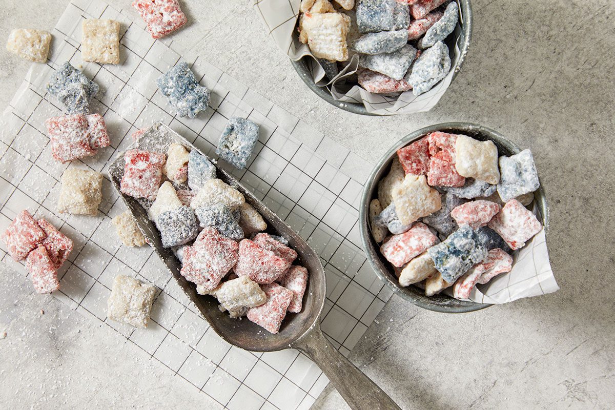 Overhead horizontal image of colorful, sugar-dusted cereal pieces arranged on a light grey surface with grid-patterned parchment paper. Red, white/tan, and blue cereal pieces are displayed in an antique-looking metal scoop, two small metal bowls, and scattered loosely around, with powdered sugar dustings visible.