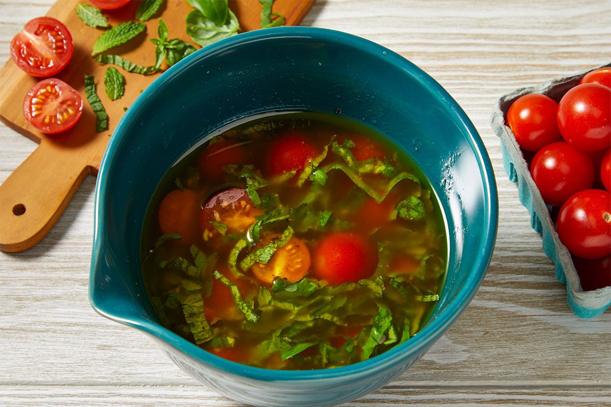 Overhead horizontal image of a blue bowl containing the vinaigrette, cherry tomatoes, julienned mint leaves, and julienned basil for Red, White & Blue Summer Salad. A carton of cherry tomatoes and a wooden cutting board with julienned mint and basil are beside the bowl.