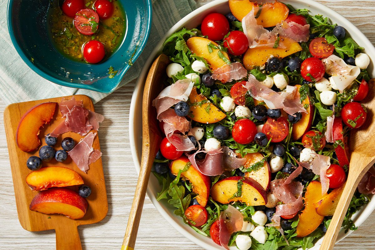 Overhead horizontal image of Red, White & Blue Summer Salad. The image shows the salad in a white bowl, tossed with wooden serving spoons, and garnished with arugula, mozzarella pearls, blueberries, peaches, tomatoes and prosciutto. A small wooden board is adjacent to the bowl, topped with peach slices, blueberries and prosciutto; a blue bowl containing the dressing sits at the upper left.