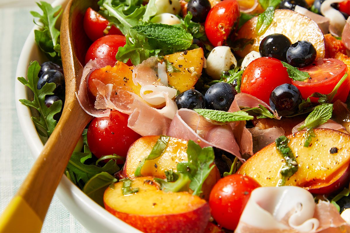 Eye Level horizontal image of Red, White & Blue Summer Salad being tossed with a wooden spoon. Visible ingredients include fresh arugula, cherry tomatoes, mozzarella cheese pearls, peaches, blueberries, prosciutto, and mint leaves.