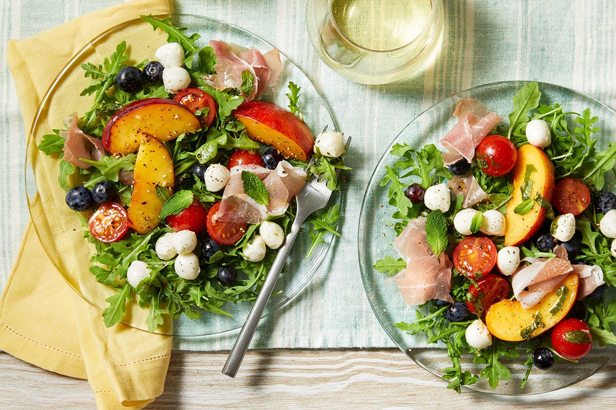 Overhead horizontal image of two clear plates filled with Red, White & Blue Summer Salad. The salad contains arugula, mozzarella cheese pearls, peaches, blueberries, tomatoes, prosciutto, and mint.
