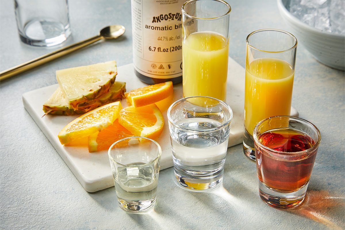 Eye Level horizontal image of various cocktail ingredients arranged on a white marble board. The setup includes pineapple wedges, orange slices, a bottle of Angostura Aromatic Bitters, along with clear and amber liquids in shot glasses, and yellow liquids in tall glasses, with a bowl of ice visible in the background. | [Original] RMS 283121 Rum Swizzle; How to; Step 1 Combine ingredients in a Collins glass (ingredient shot)
