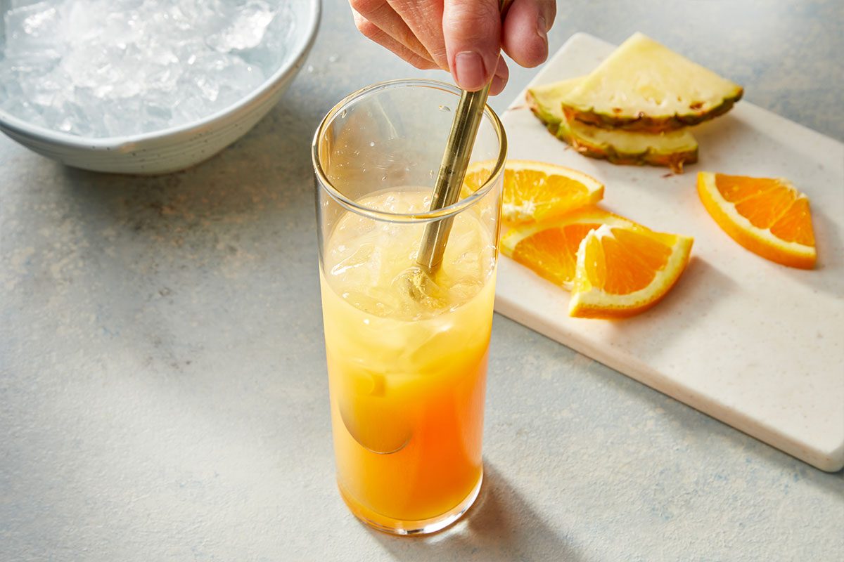 Eye Level horizontal image of a hand stirring a vibrant orange and yellow beverage in a tall glass filled with ice. Fresh orange slices and pineapple wedges are arranged on a white cutting board next to the drink, with a bowl of ice visible in the background. | [Original] RMS 283121 Rum Swizzle; How to; Step 2; Add crushed ice until the glass is 3/4 full. Swizzle with a swizzle stick or bar spoon.