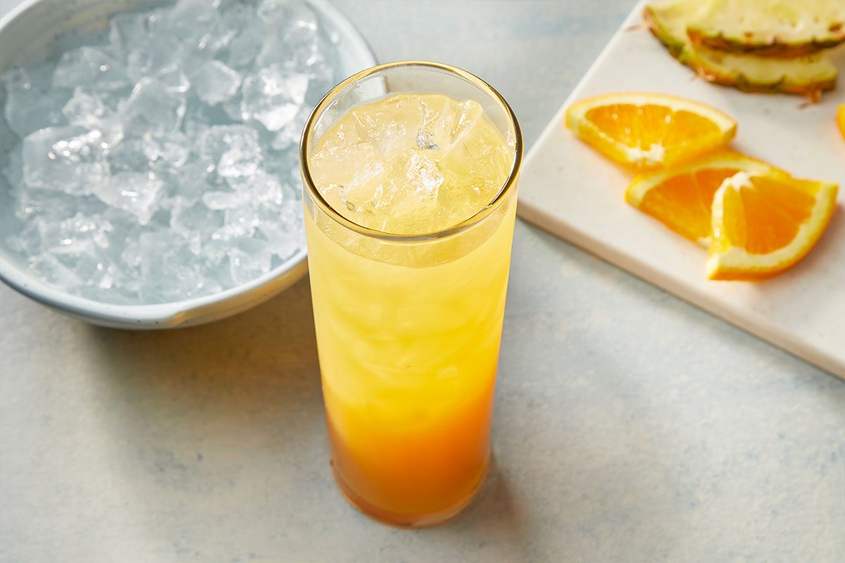 Eye Level horizontal image of a tall glass filled with a refreshing yellow and orange layered drink with ice cubes. A bowl of ice is visible in the background, along with orange slices and pineapple slices on a white cutting board. | [Original] RMS 283121 Rum Swizzle; How to; Step 3; Top with additional crushed ice as needed to fill the glass