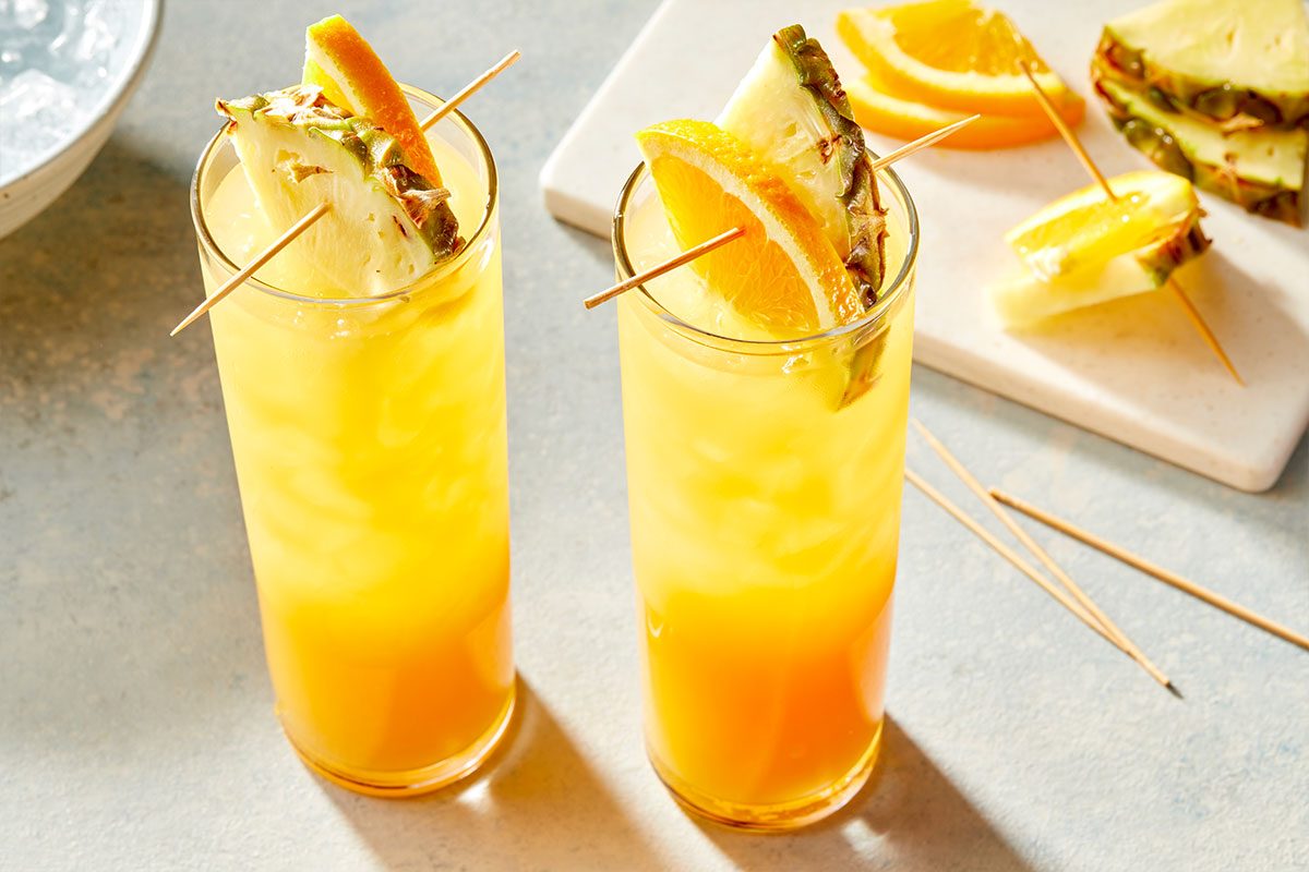 Eye Level horizontal image of two tall glasses filled with a layered yellow and orange drink, each garnished with a fresh pineapple slice and an orange wedge on a skewer. In the background, additional pineapple and orange slices are arranged on a white board, with extra skewers and a bowl of ice partially visible.