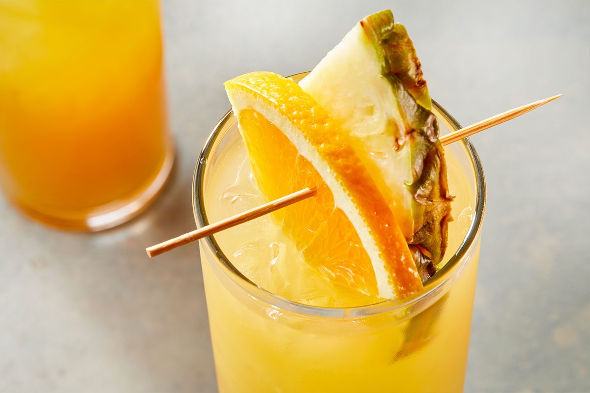 Eye Level horizontal image of a vibrant yellow drink in a clear glass filled with ice. The glass is garnished with a fresh orange slice and a pineapple wedge, held together by a wooden skewer. Another glass, out of focus, is visible in the background.