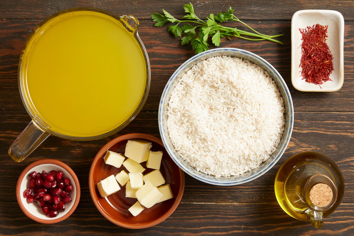 Overhead horizontal image of various raw ingredients displayed on a dark wooden surface. A clear glass measuring cup holds a generous amount of yellow broth, positioned near a bowl brimming with white rice, a dish of vibrant red saffron threads, and fresh green parsley. Other visible items include small cubes of butter, ruby-red pomegranate seeds, and a small bottle of olive oil.