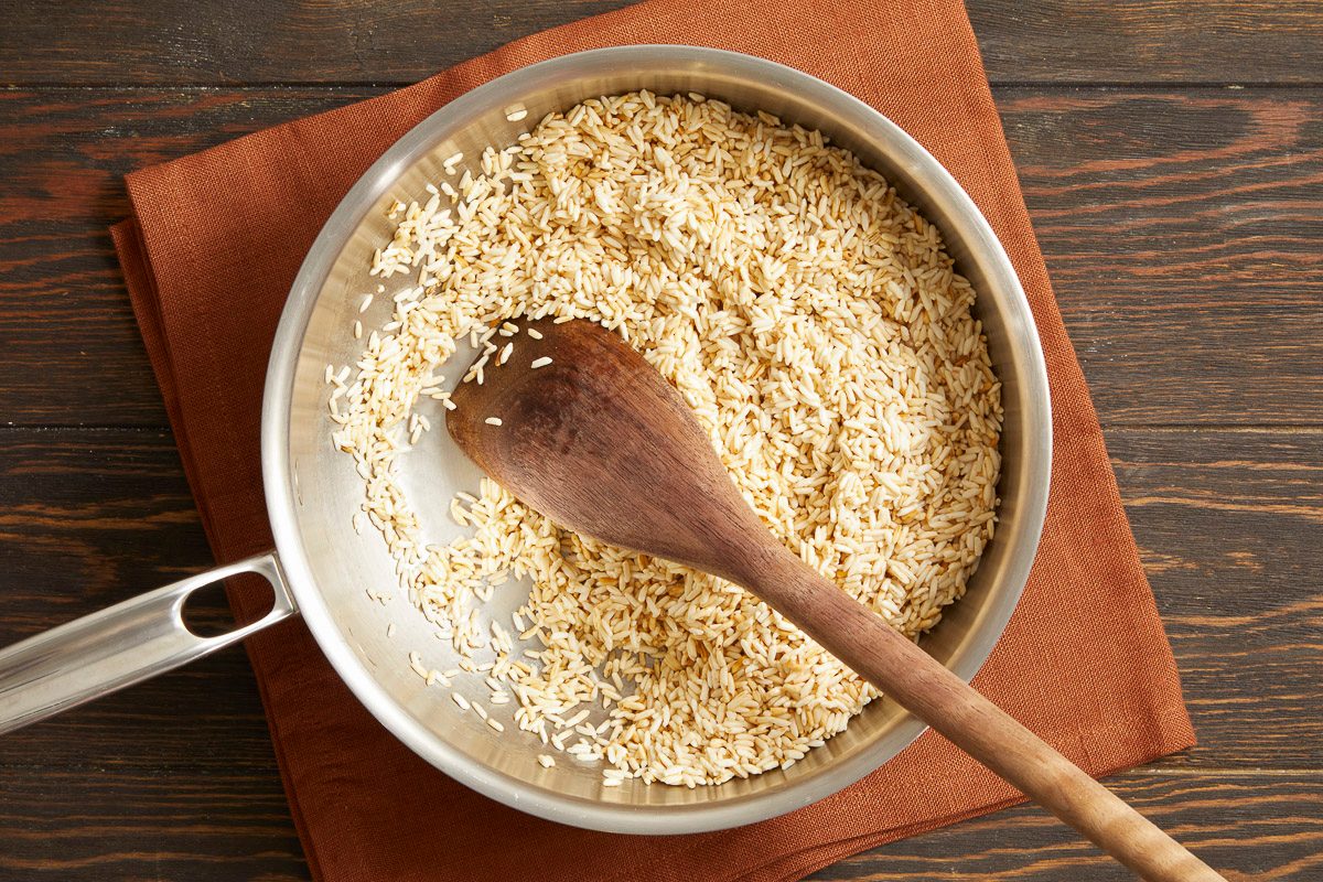 Overhead horizontal image of uncooked rice in a stainless steel pan, with a wooden spoon resting in the grains. The pan sits on a brown napkin over a dark wooden surface.