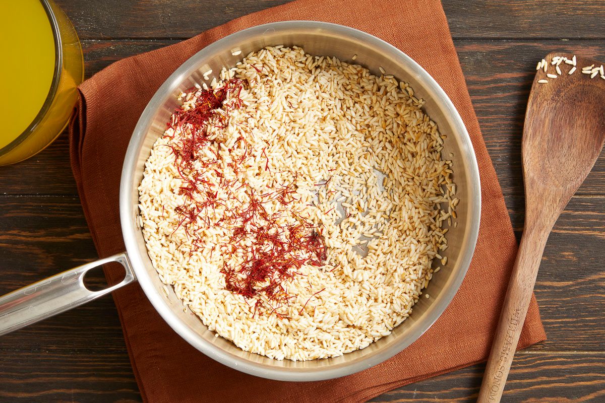 Overhead horizontal image of saffron threads and rice in a silver pan. A wooden spoon is placed next to the pan on a brown cloth, with a glass bowl containing yellow broth in the background.