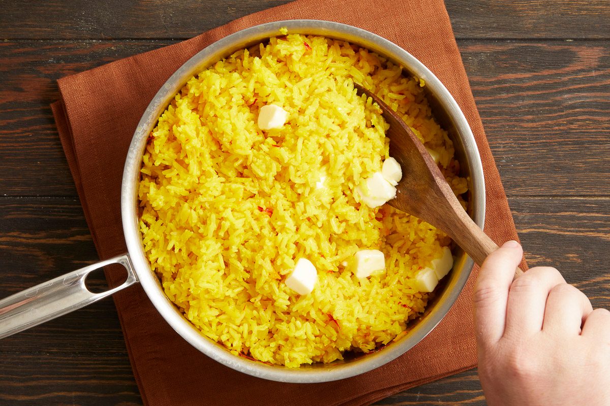Overhead horizontal image of a hand stirring vibrant yellow rice with pats of butter melting into it, using a wooden spoon in a silver pan. The rice appears to be saffron-infused, resting on a brown napkin.