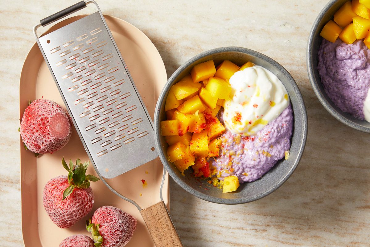 Overhead horizontal image of frozen strawberries on a tray next to a metal grater, showing some grated berry. In the foreground, a bowl of purple chia pudding is topped with diced mango and a dollop of cream, garnished with grated strawberry.