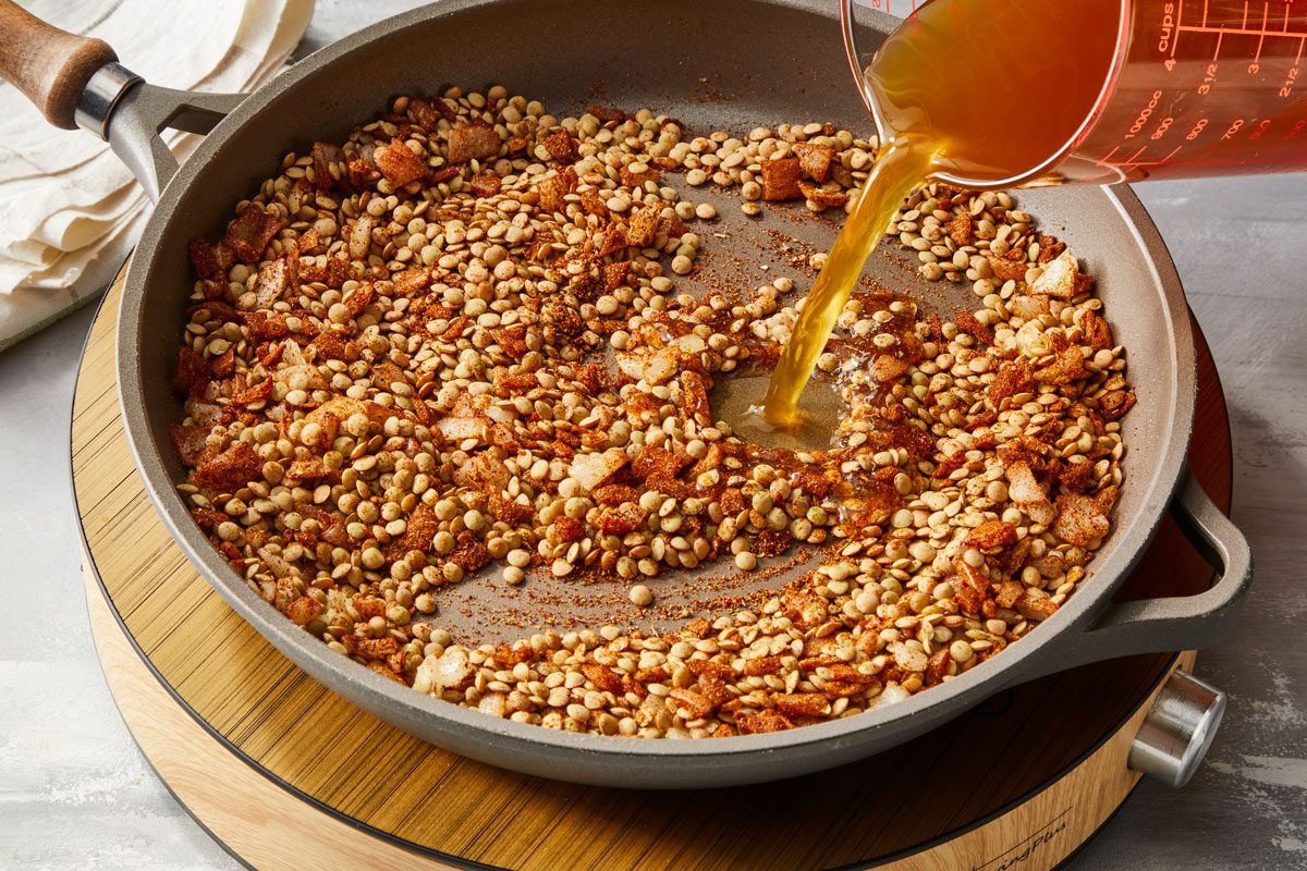 Overhead view of the preparation of lentil taco filling. Vegetable broth is being poured into a pan containing lentils, chopped onion, and spices.