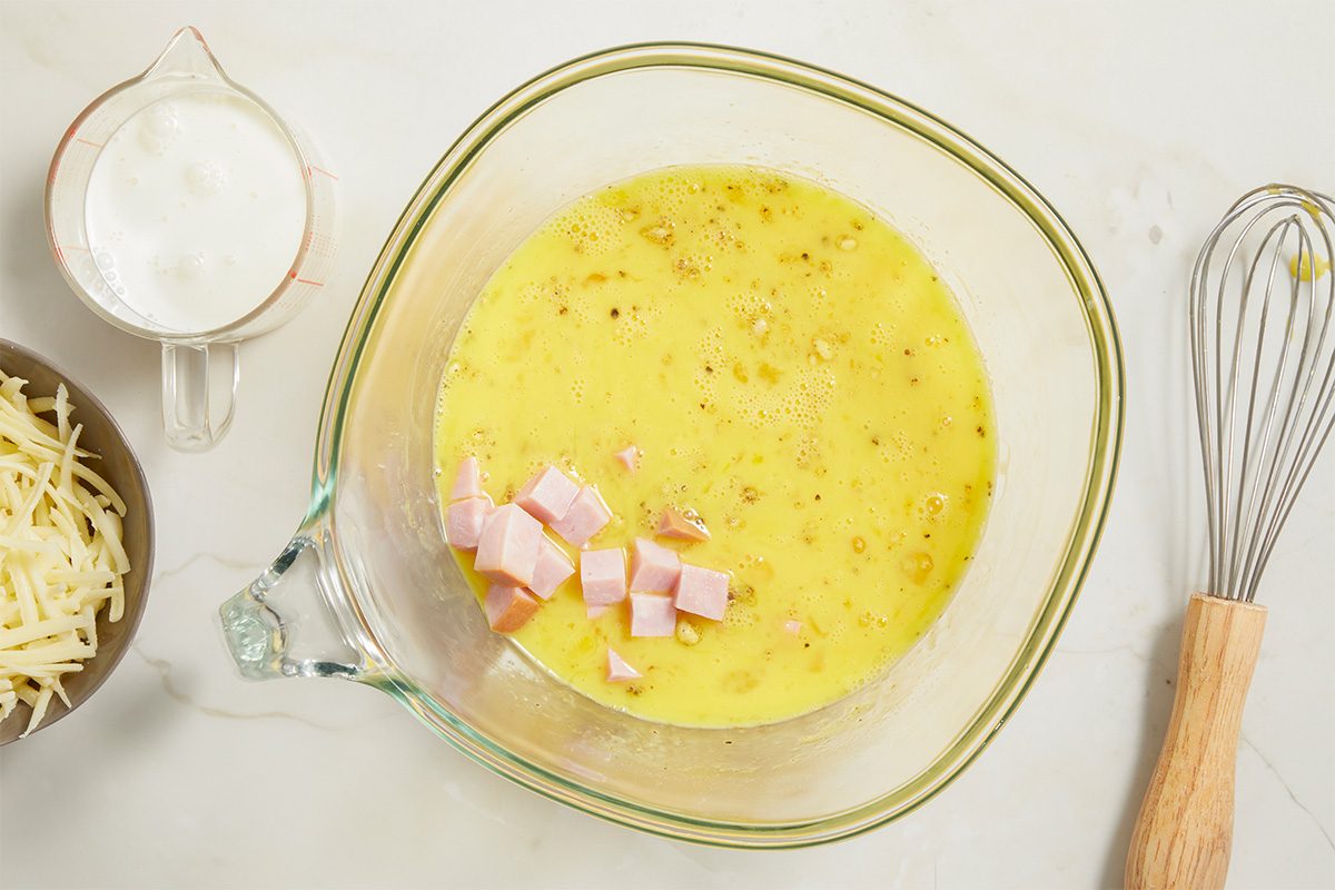 Overhead horizontal image of Slow-Cooker Ham & Gruyere Quiche, showing the step of adding cubed ham to a clear glass mixing bowl that also contains the whisked egg mixture. In a separate bowl are shredded Gruyere cheese and in a glass measuring cup is the heavy whipping cream.