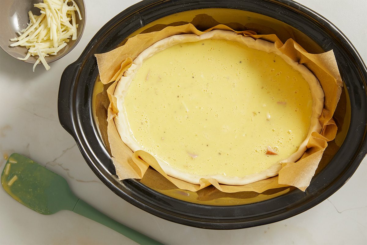 Overhead horizontal image of a slow cooker with a quiche being prepared. The pie crust is lined with parchment paper and filled with an egg and ham mixture. Gruyere cheese is in a bowl to the side.