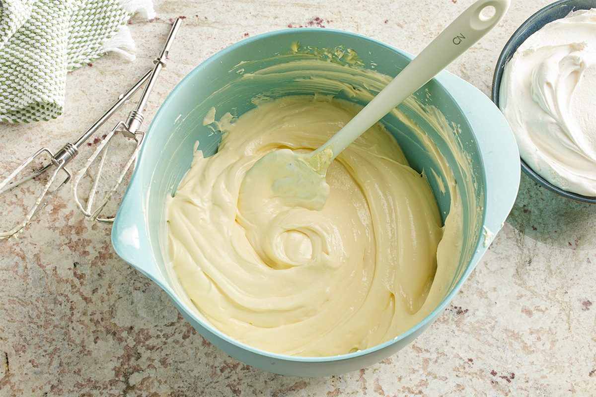 Overhead horizontal image of a pale yellow creamy mixture being prepared in a light blue mixing bowl, with a silicone spatula resting inside. To the left, electric mixer beaters and a green and white checkered towel are visible, alongside a bowl of thawed whipped topping.