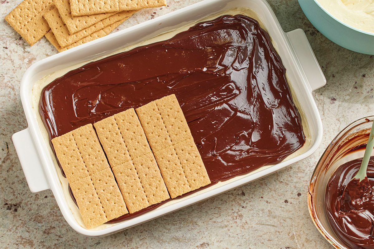 Overhead horizontal image of a white baking dish containing layers for Horchata Icebox Cake. Whole graham crackers are being placed on top of a dark chocolate layer, which sits over a lighter creamy layer. More whole graham crackers are scattered on the counter.
