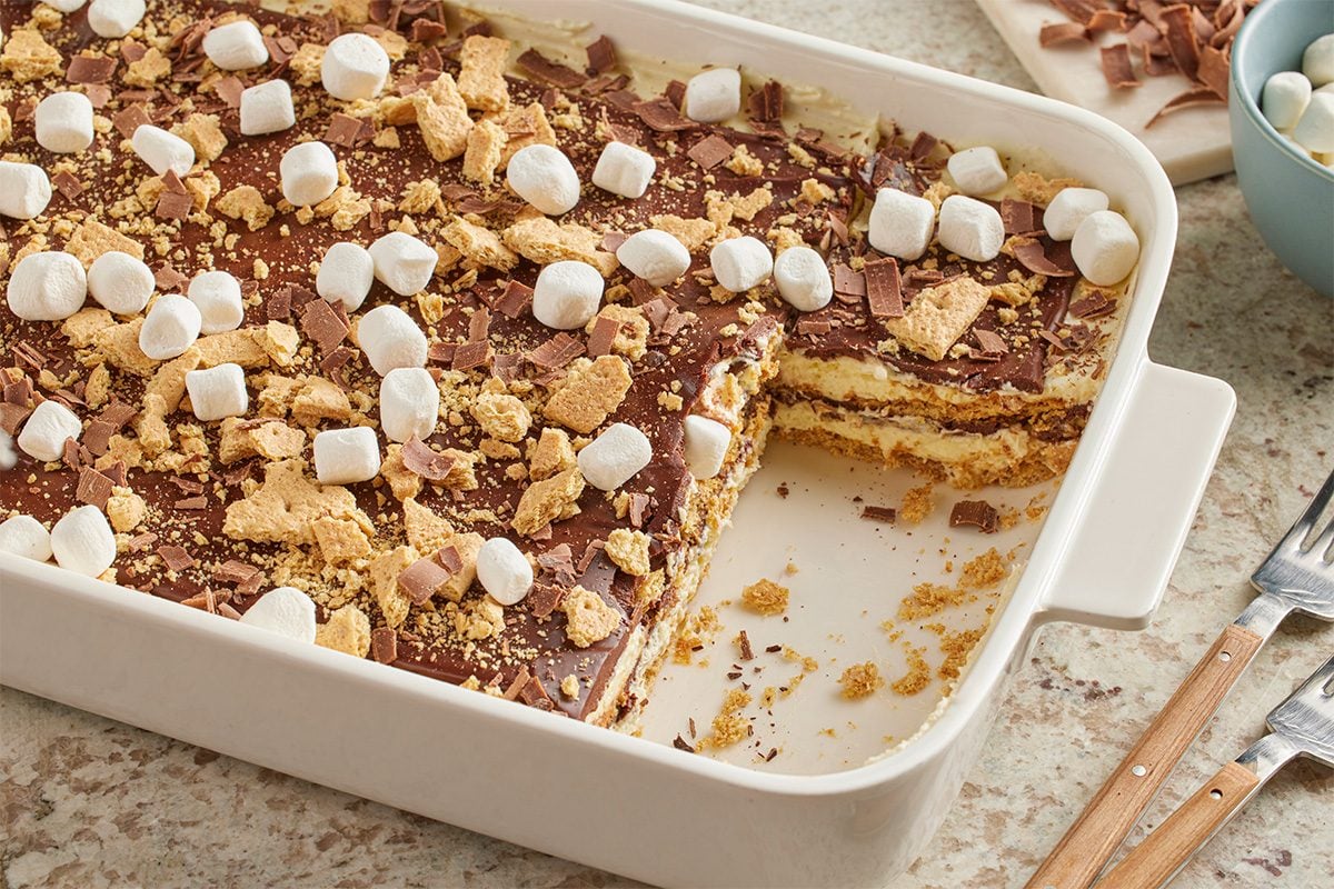 Eye level horizontal image of a Horchata Icebox Cake in a white rectangular baking dish with a slice removed. The layered dessert features crushed graham crackers, pudding, and is topped with chocolate shavings, additional crushed graham crackers, and mini marshmallows. A bowl of marshmallows and more chocolate shavings are visible in the background.