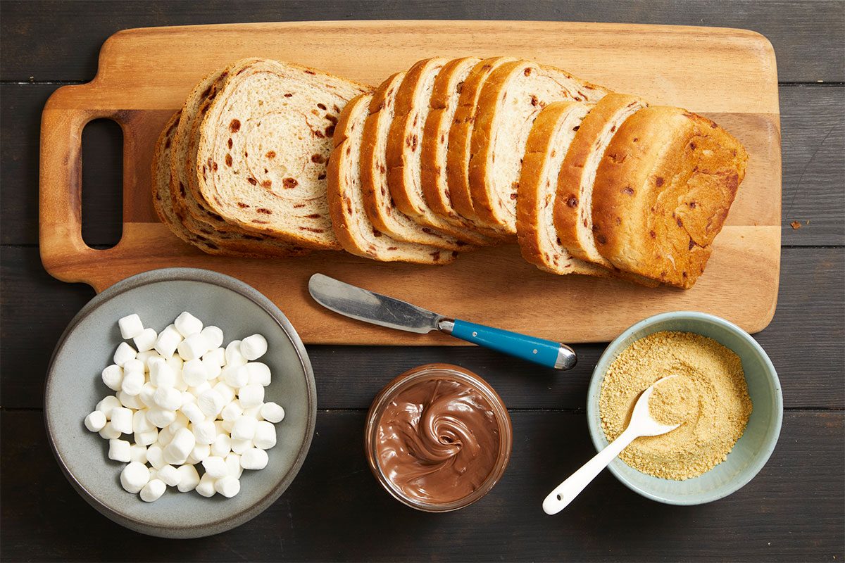 Overhead horizontal image of ingredients laid out on a dark wooden surface. A loaf of sliced cinnamon bread rests on a wooden cutting board, alongside bowls of miniature marshmallows and crushed graham crackers, and an open jar of Nutella. A knife and spoon are also visible.