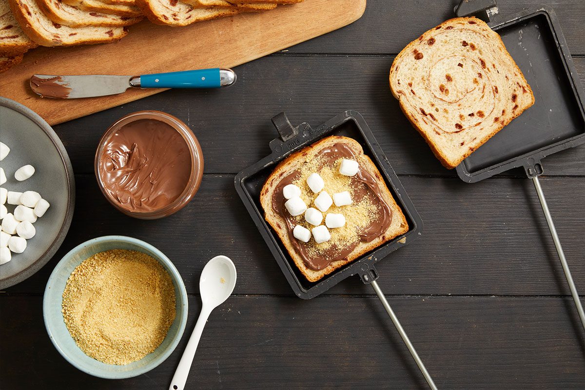 Overhead horizontal image of ingredients being assembled for S'more Pudgy Pie in a pie iron. A slice of cinnamon bread spread with Nutella, graham cracker crumbs, and miniature marshmallows is in a pie iron, with another slice of cinnamon bread next to it. A jar of Nutella, a bowl of miniature marshmallows, and a bowl of crushed graham crackers are also visible on a dark wooden surface.