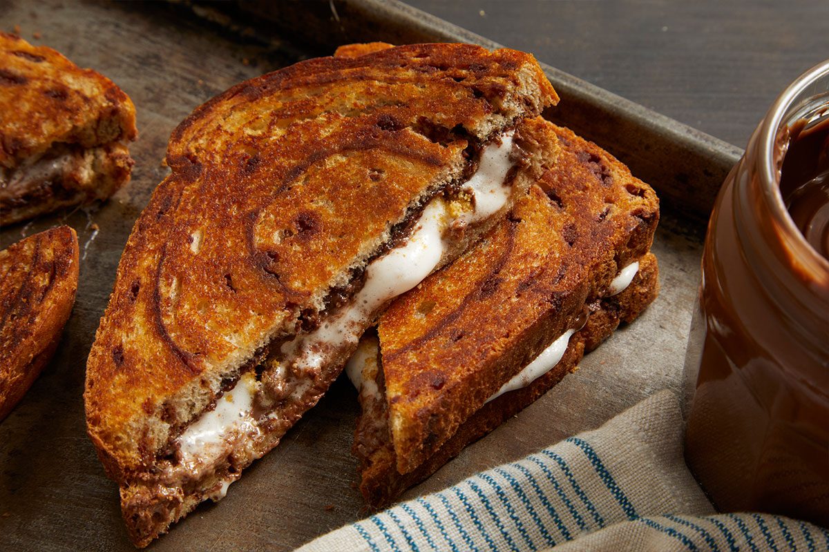 Eye Level horizontal image of a S'more Pudgy Pie, cut in half, revealing melted marshmallow and Nutella chocolate spread. The golden-brown sandwich rests on a dark metal tray, with a jar of Nutella and a striped cloth napkin partially visible in the background.