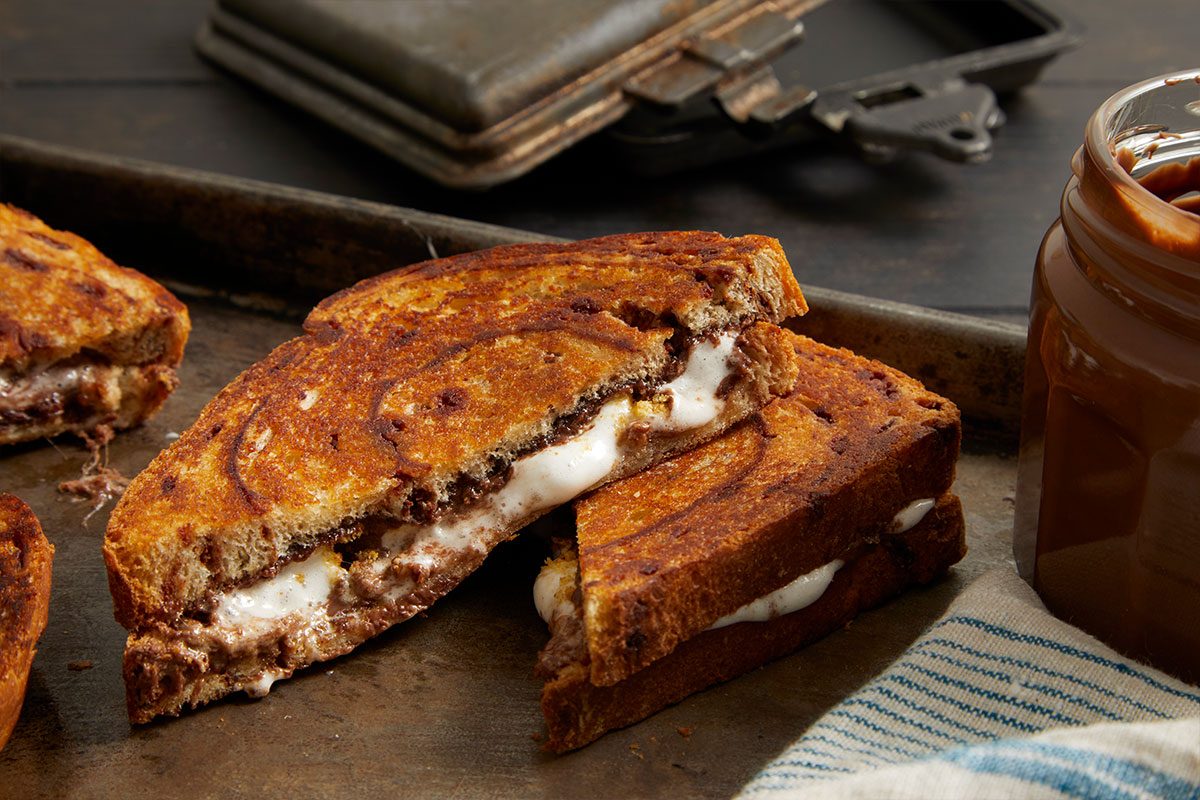 Eye Level horizontal image of two S'more Pudgy Pies, cut in half and showing the gooey, melted marshmallow and Nutella filling. The toasted cinnamon bread sandwiches sit on a dark baking sheet, with a jar of Nutella and a striped cloth in the background.