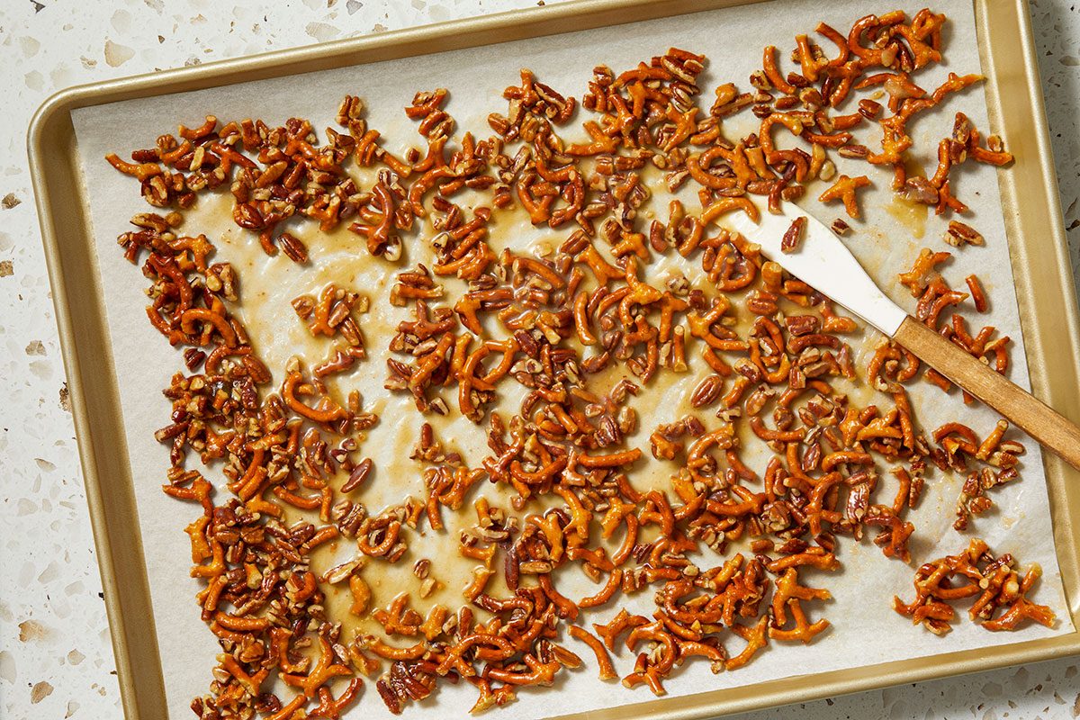 Overhead horizontal image of mini pretzels and chopped pecans coated in a shiny, golden glaze, spread out on parchment paper on a gold-toned baking sheet. A white and wooden spatula is partially visible on the right side.
