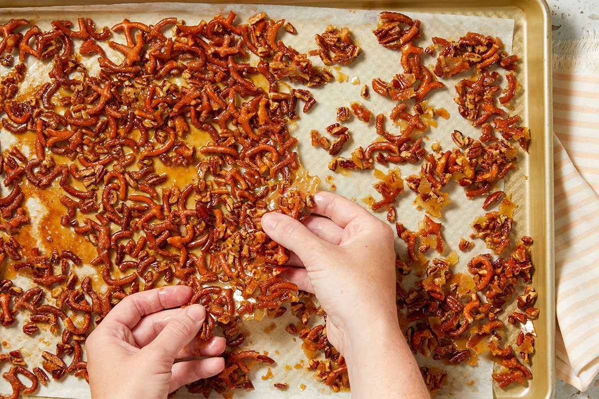 Overhead horizontal image of two hands breaking apart clusters of pretzels and pecans coated in a sticky caramel glaze. The mixture is spread on a parchment-lined baking sheet.