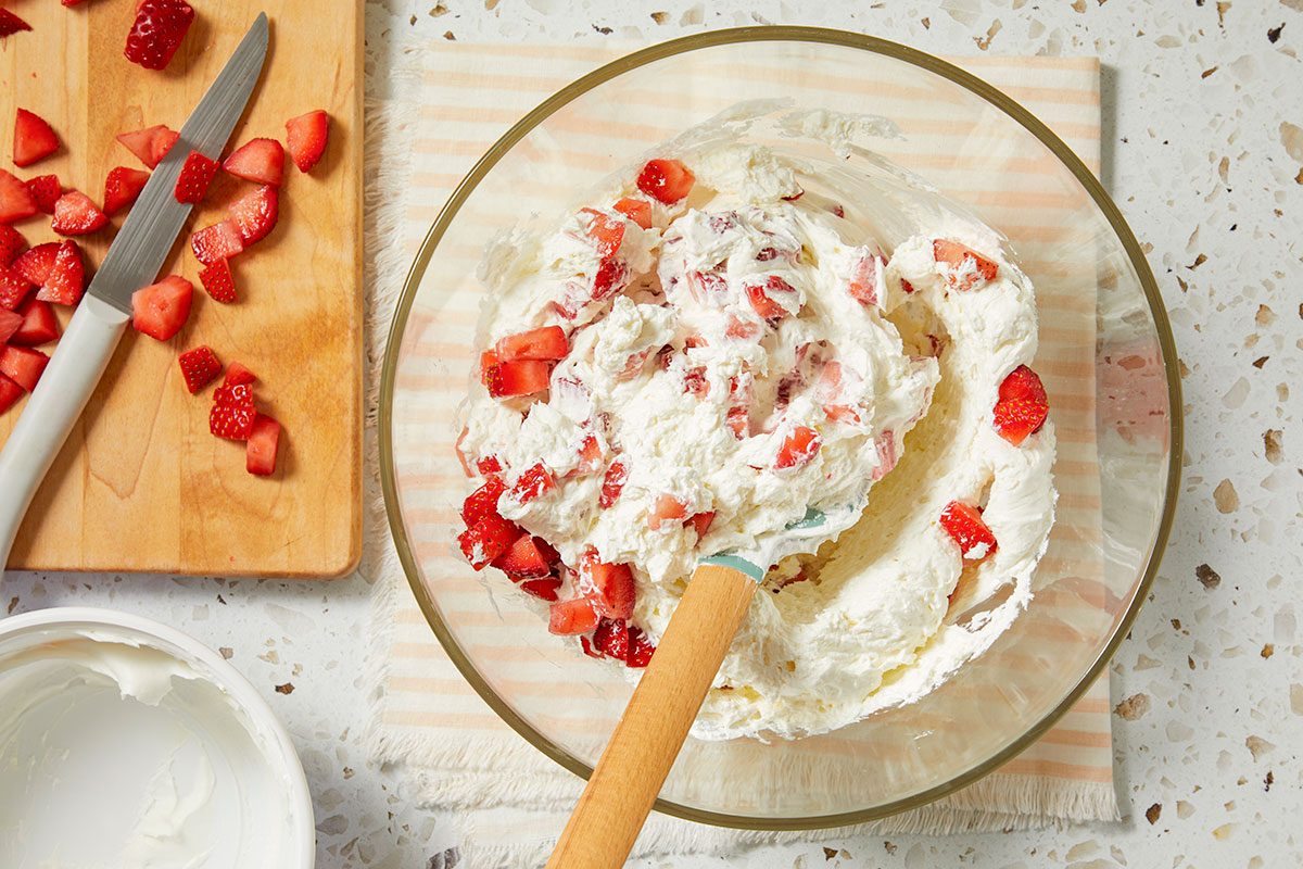 Overhead horizontal image of diced strawberries being folded into a creamy white mixture in a clear glass bowl. A spatula is actively mixing the ingredients, while a wooden cutting board with more diced strawberries and a knife is visible nearby. A white container of cream sits to the lower left.